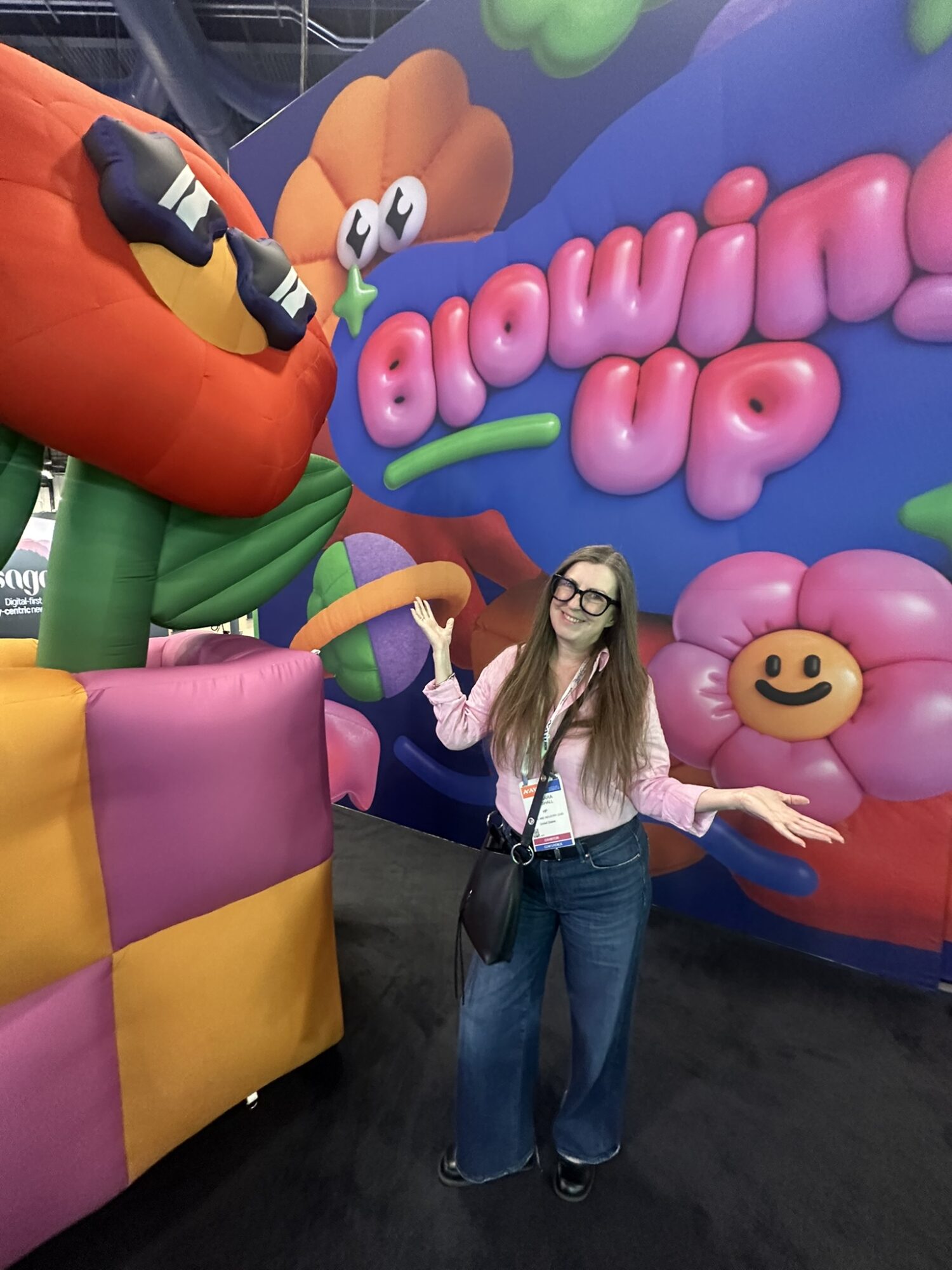 Woman with glasses and long hair stands in front of colorful inflatables and a sign that says 'Blowout Up'.