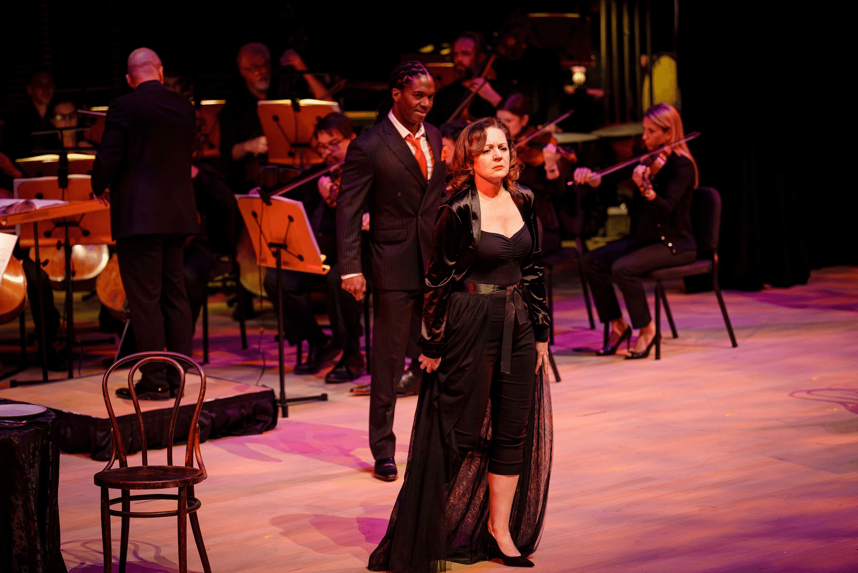 Two performers stand on stage with an orchestra in the background, one woman in a black dress and one man in a suit.