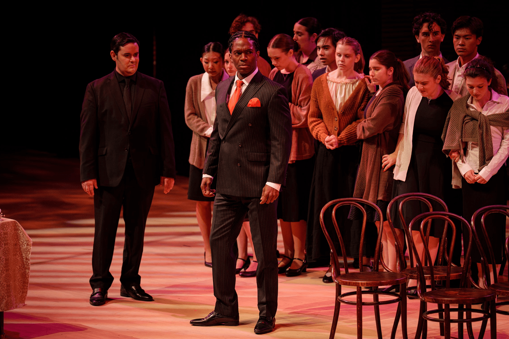 Group of people standing on stage, some seated chairs visible, dark background, formal attire, diverse group, serious expressions.
