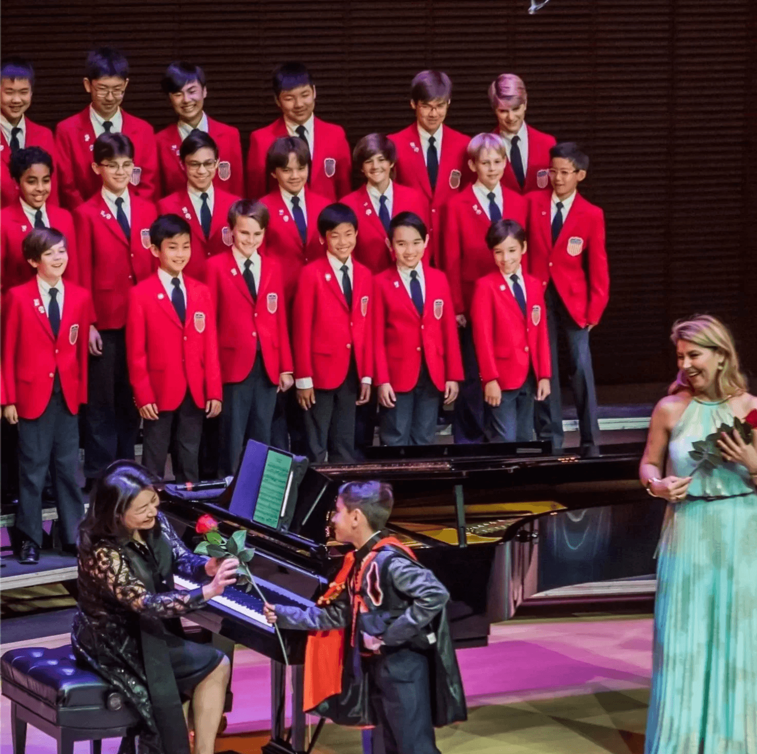 Group of students in red blazers on stage, two children at piano, woman in colorful dress holding papers, stage setting.