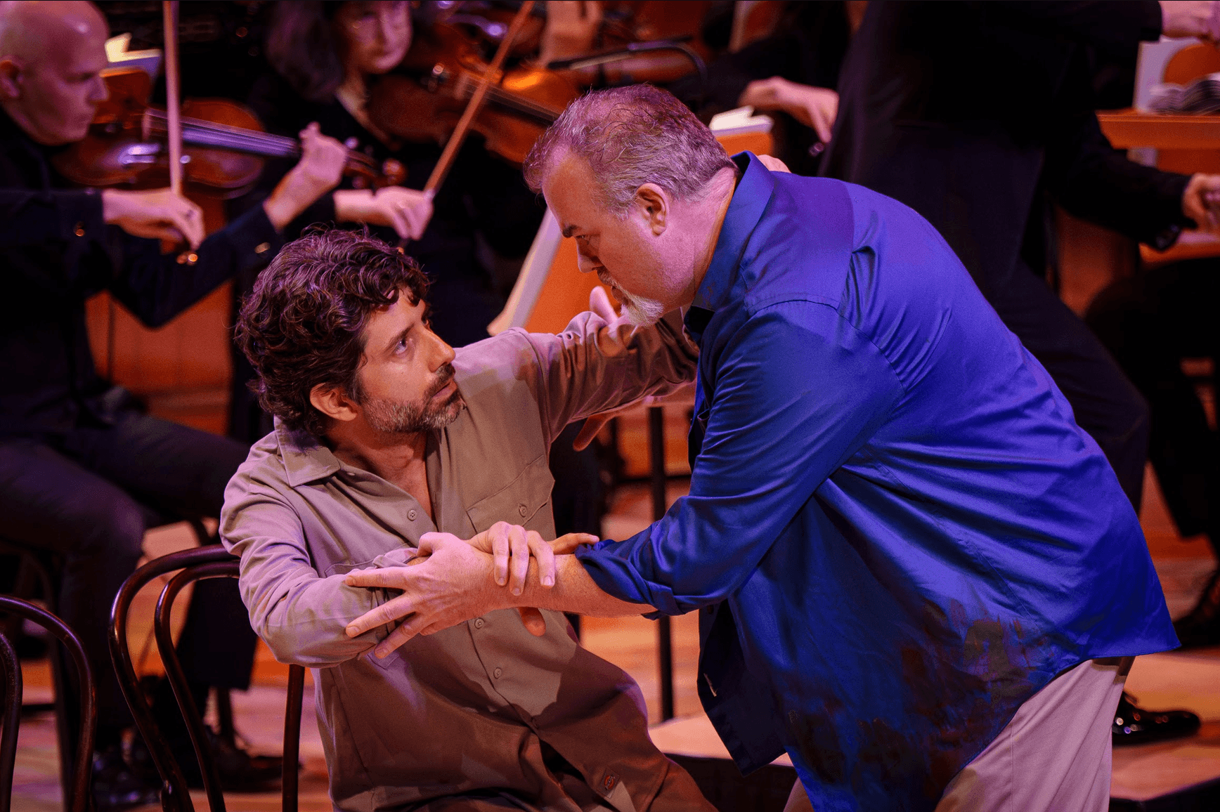 Two men interact on stage with an orchestra in the background, one seated and the other standing, holding hands.