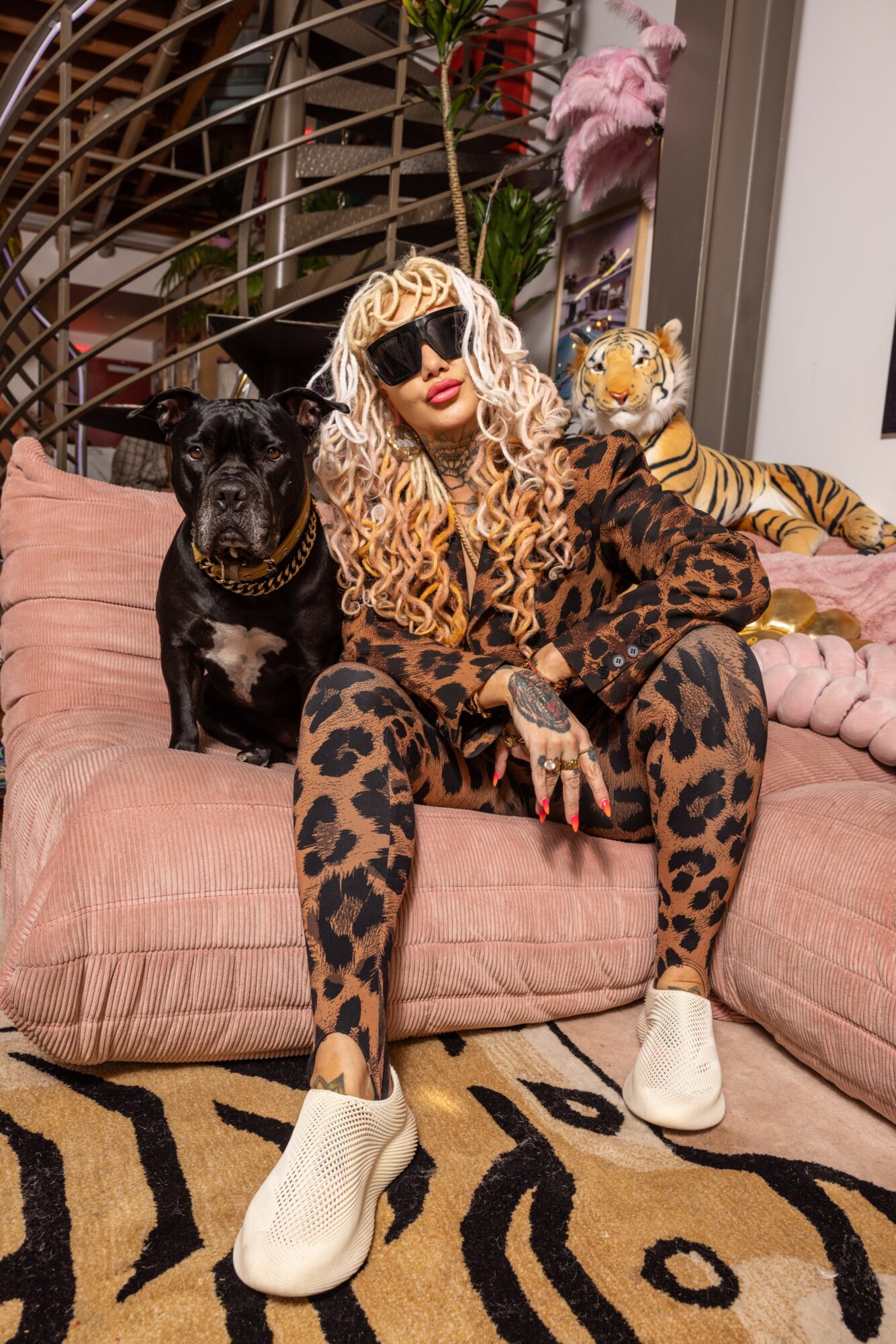 Person with long curly blonde hair, sunglasses, leopard print outfit, sitting on pink sofa with a black dog and a stuffed tiger, in a room with plants and zebra-patterned rug.