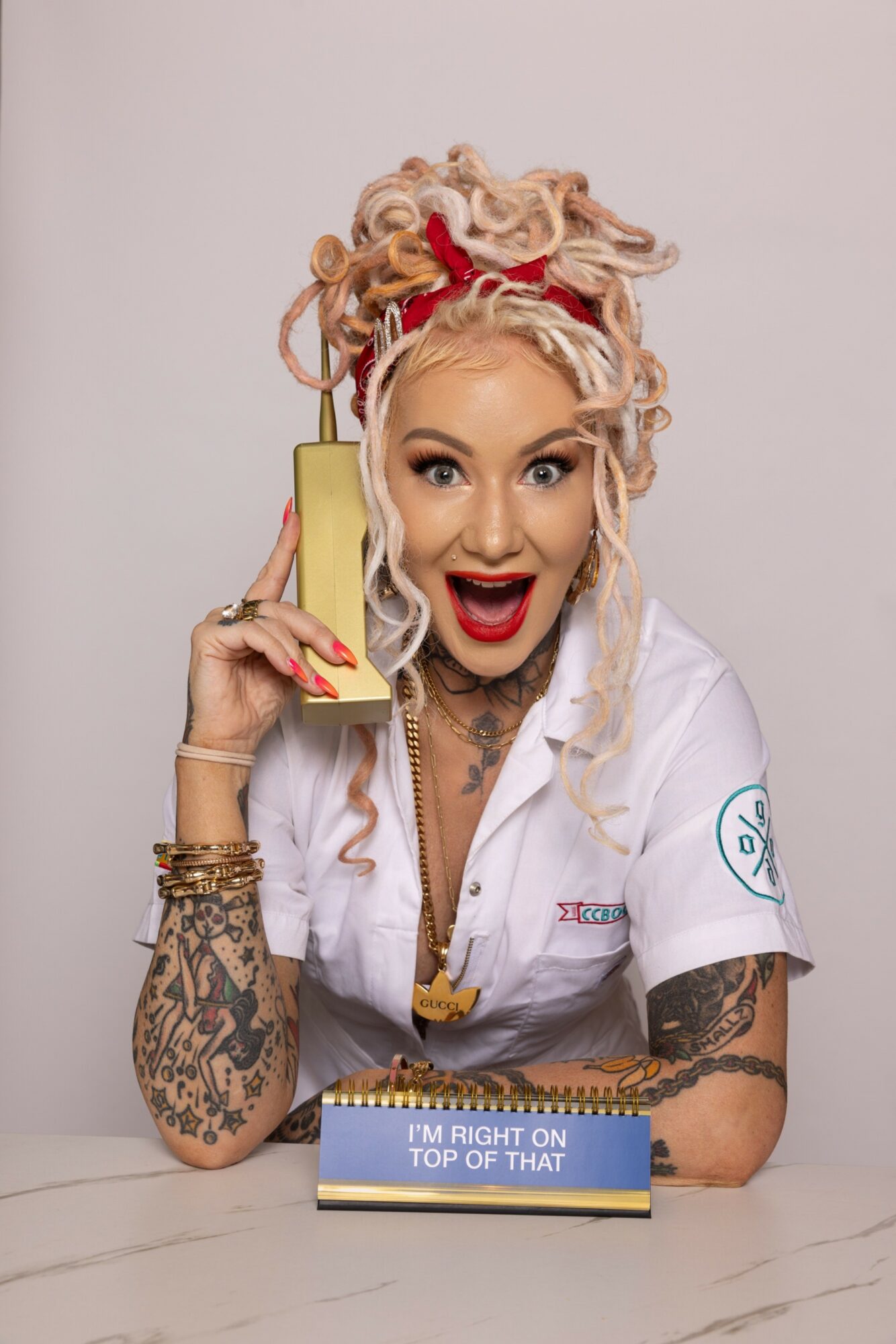 Woman with curly blonde hair, red headband, and tattoos, smiling and talking on a yellow phone, sitting at a table.