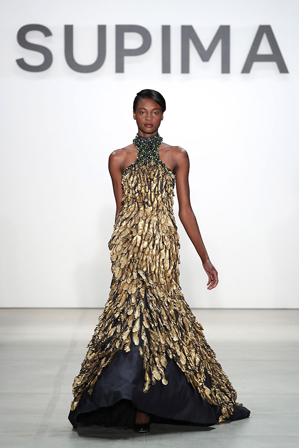 Model walking on runway in a gold and black textured gown with high neck, against white background with 'SUPIMA' text.