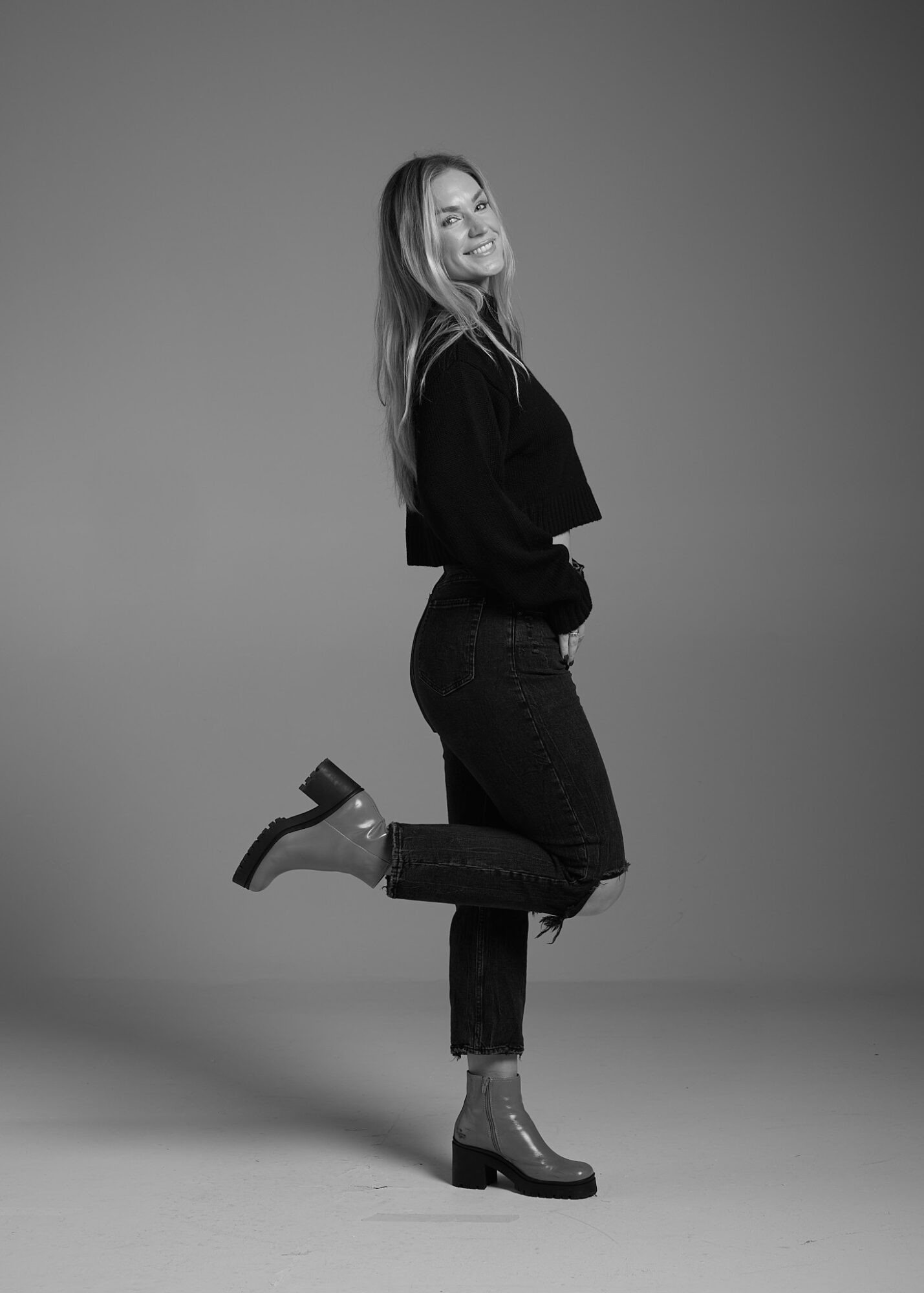 Woman smiling, standing with one leg raised, wearing a black top, jeans, and high-heeled boots, against a plain background.