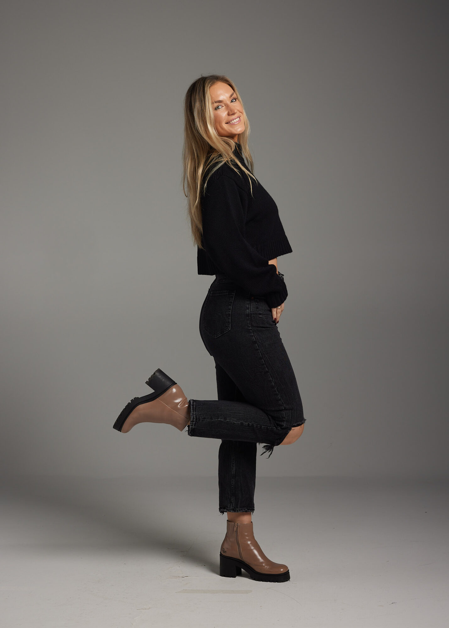 Woman with long hair smiling, standing on one leg with the other bent, wearing a black top, dark jeans, and high-heeled boots.
