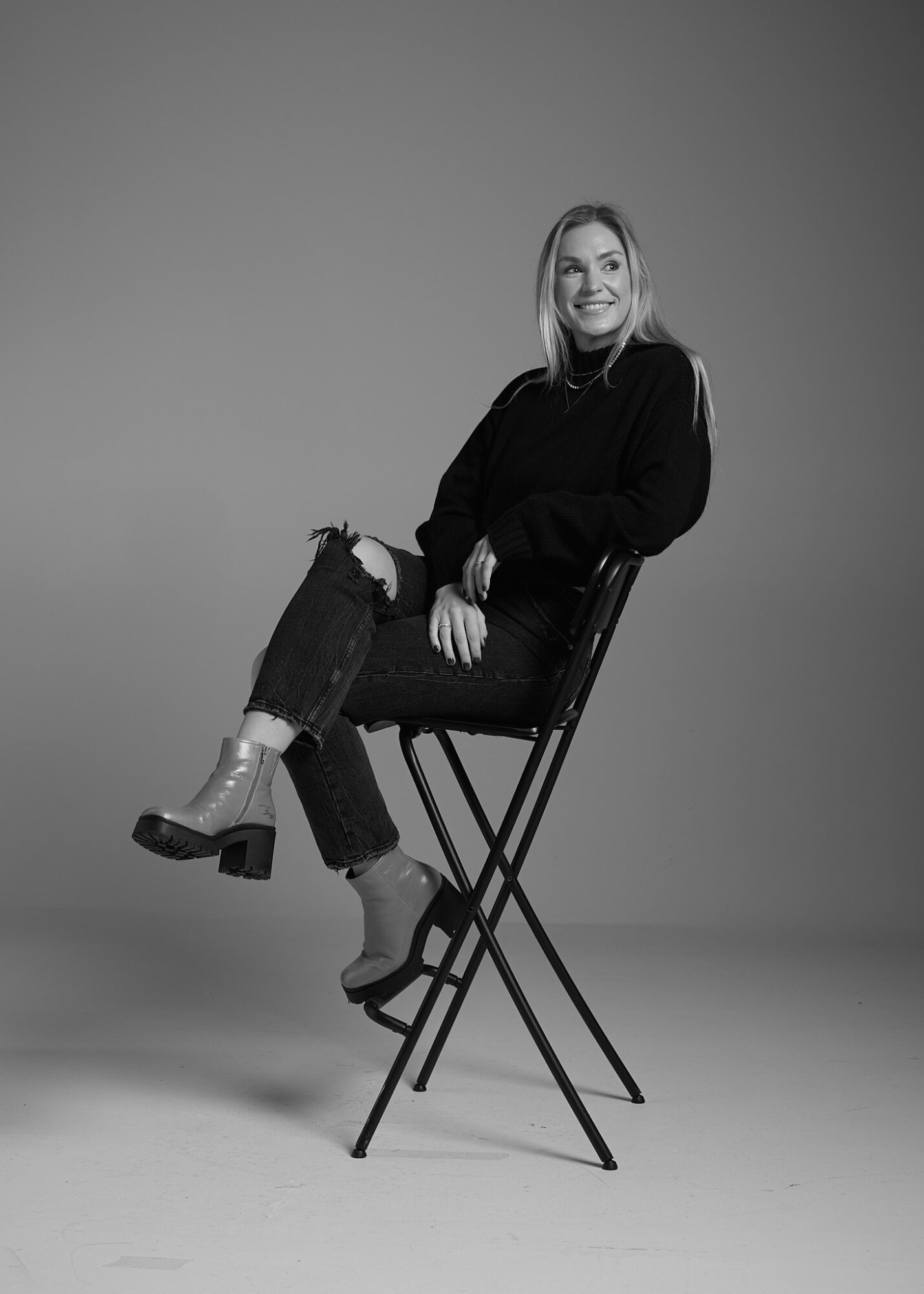 Woman sitting on a chair with crossed legs, smiling, in a studio setting, black and white photo.