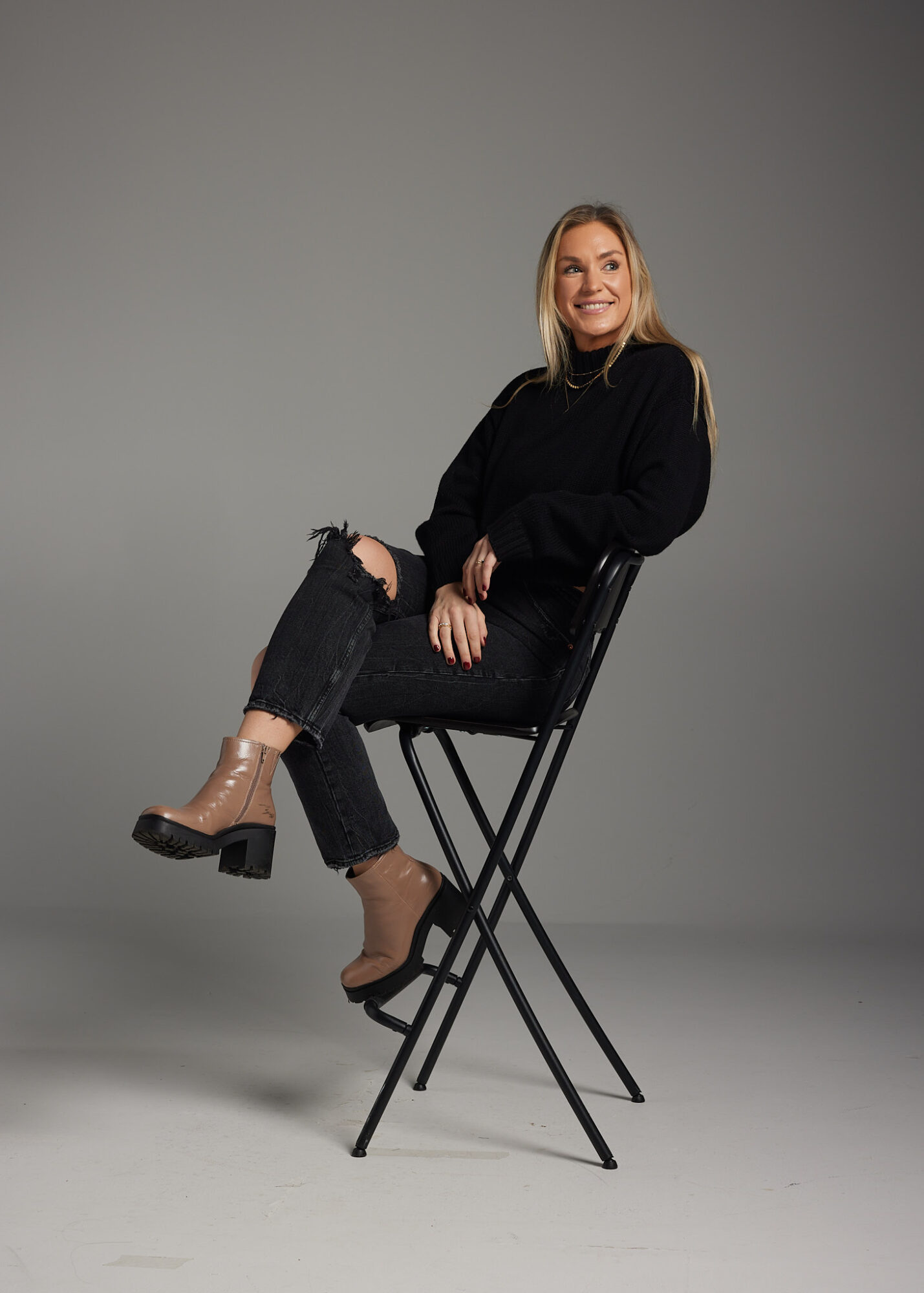 Woman sitting on a tall black chair, smiling, wearing a black sweater, ripped jeans, and beige ankle boots, against a plain gray background.