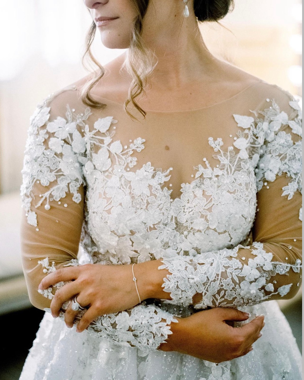Close-up of a woman in a wedding dress with floral lace details on the bodice and sleeves, hands crossed in front.