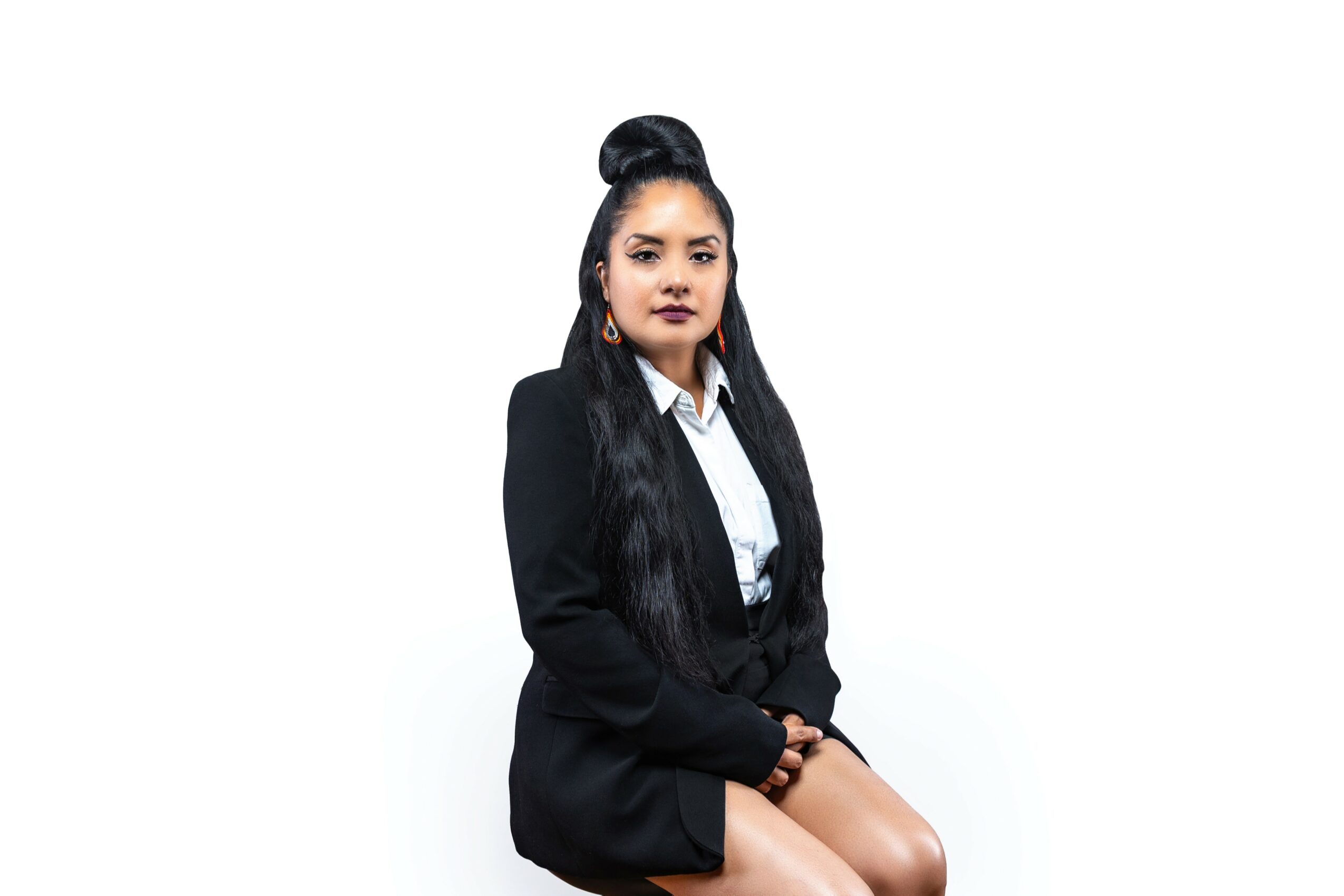 Young woman with long dark hair in a bun, wearing a black blazer and white shirt, sitting against a white background.