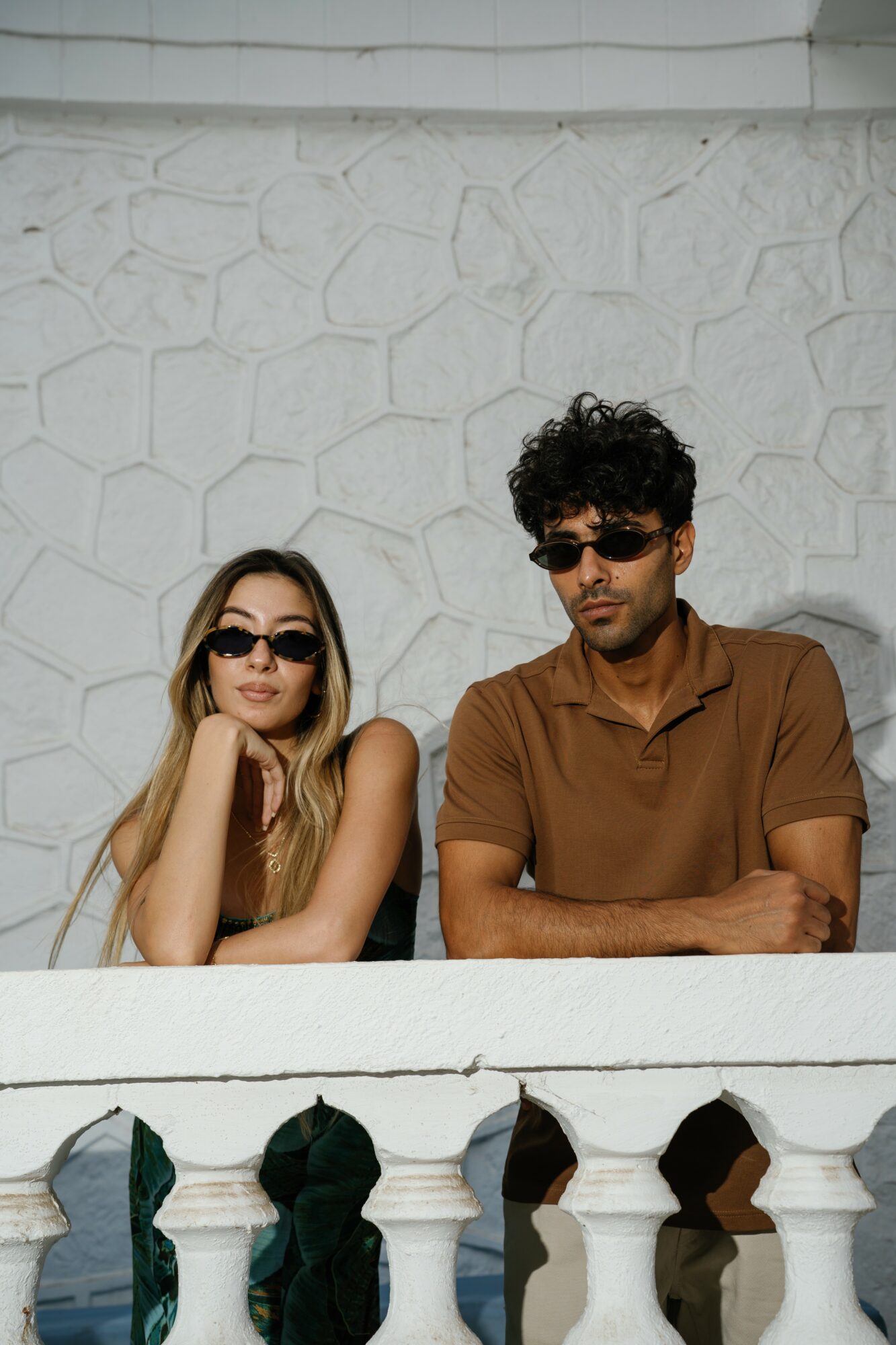 Two people with sunglasses sit against a textured wall, one resting her chin on her hand, the other with arms crossed.