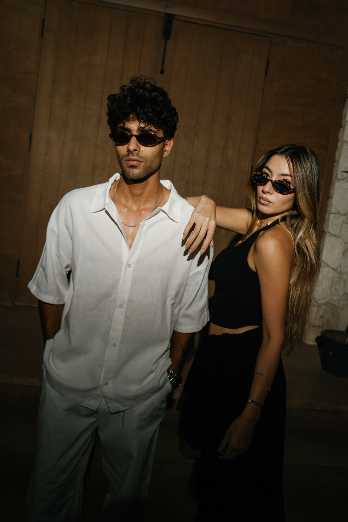 A man and woman wearing sunglasses pose indoors against a wooden background.