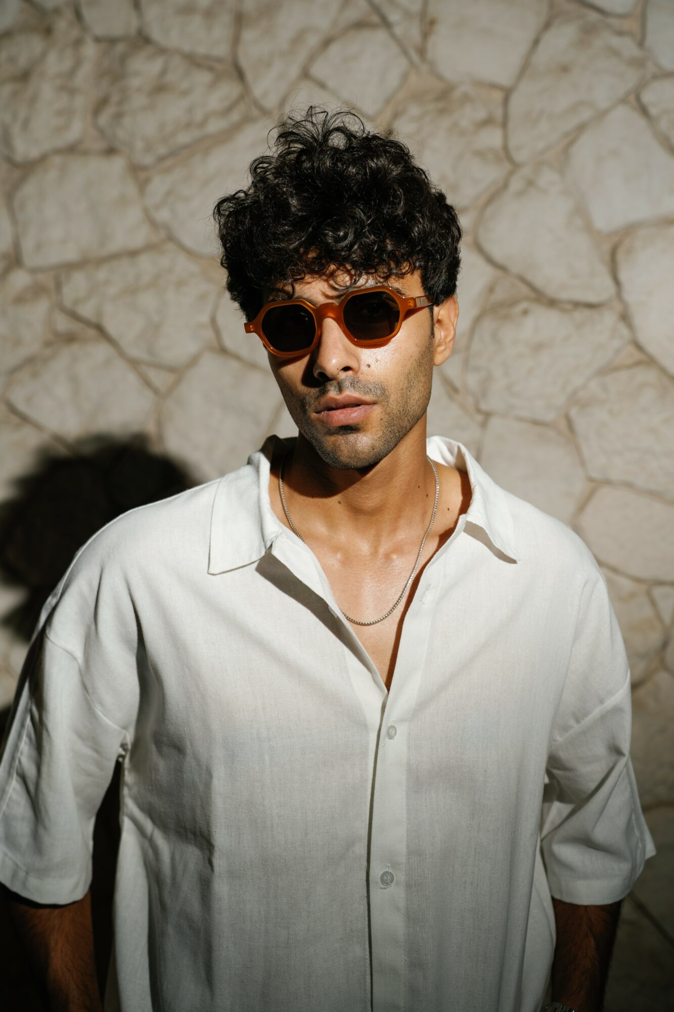 Young man with curly hair wearing sunglasses and a white shirt, standing against a stone wall.