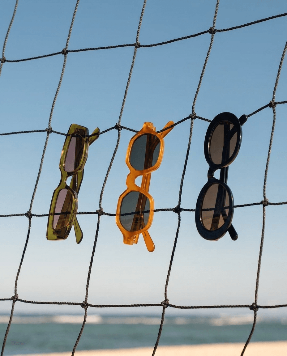 Three pairs of sunglasses hanging on a net against a blue sky and ocean background.
