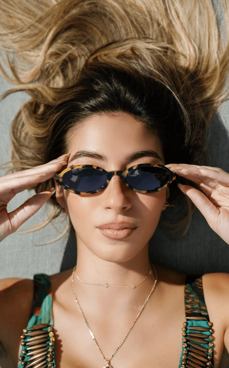 Woman with long hair wearing sunglasses and a green top, lying on a surface, holding sunglasses, with a neutral background.