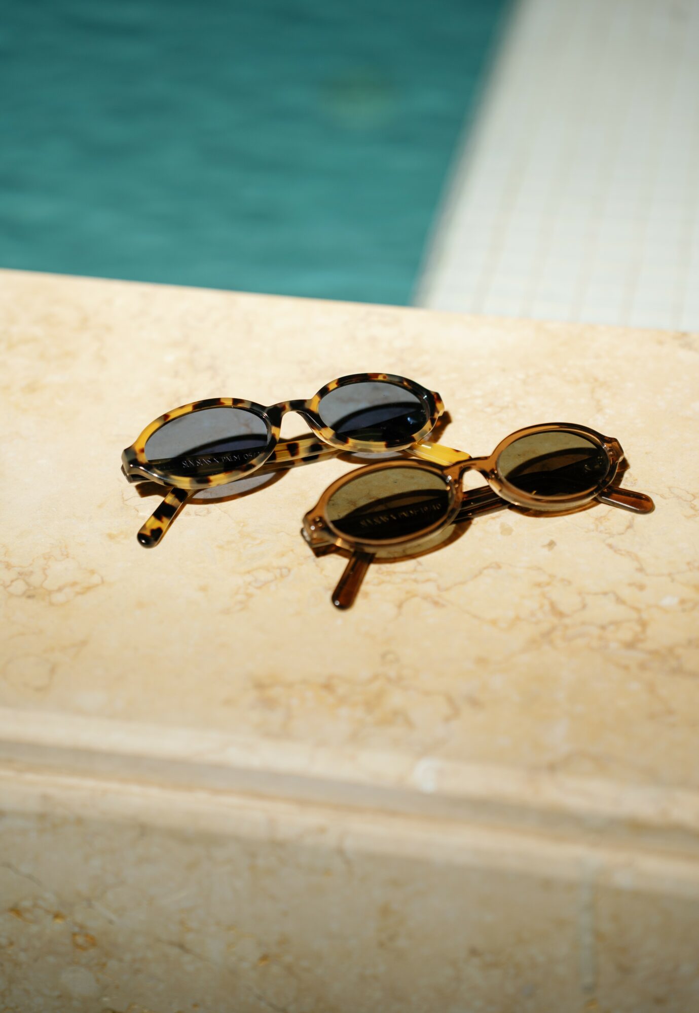 Two pairs of sunglasses with dark lenses and tortoiseshell frames resting on a beige stone surface near a pool.