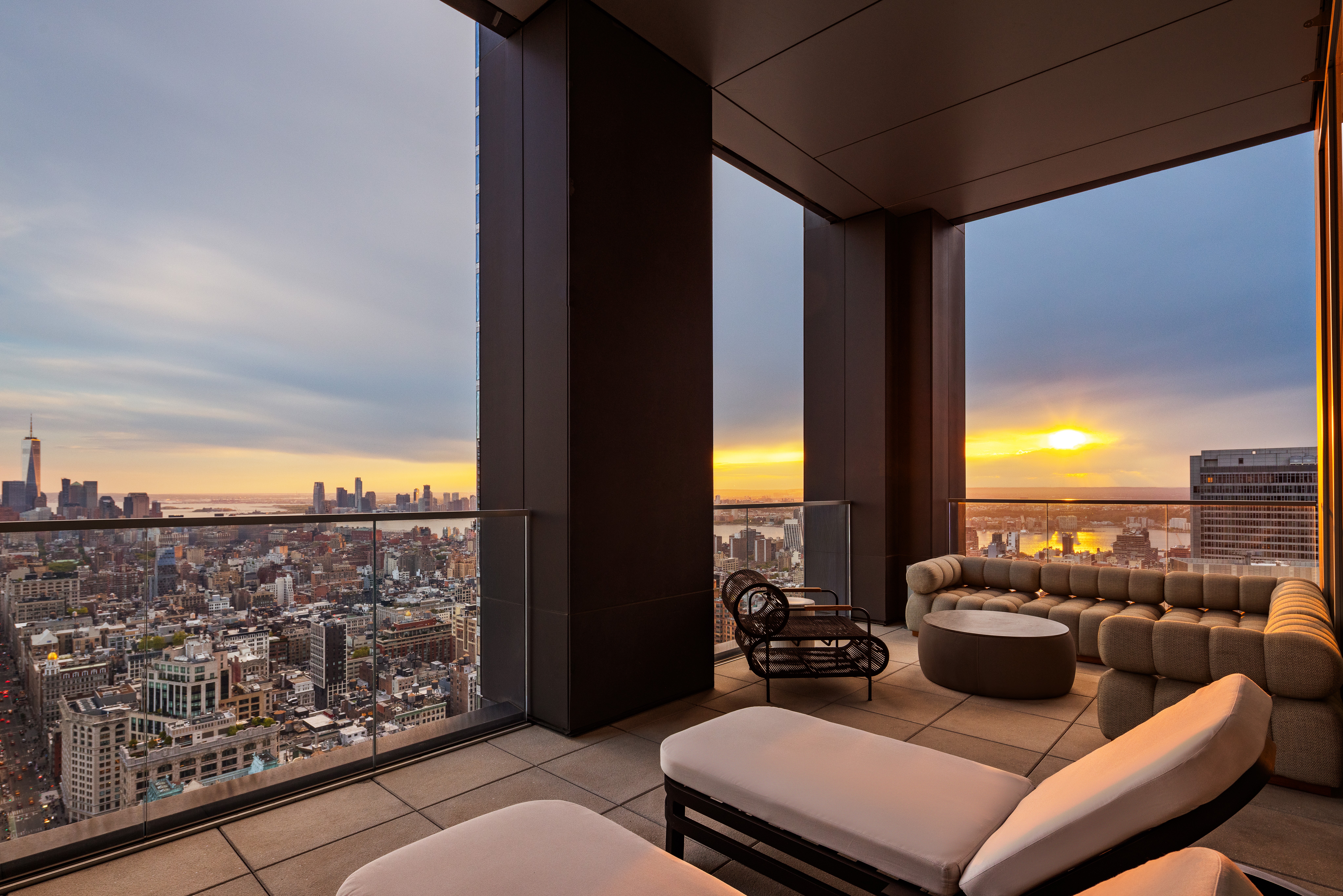 Interior of a high-rise apartment with large windows showing a city skyline at sunset, modern furniture including a sofa and chairs.