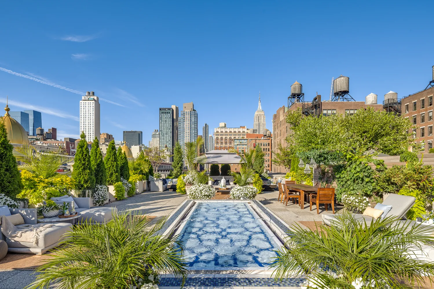 Rooftop garden with a rectangular pool, lounge chairs, tables, and city skyline in the background, with trees and greenery.