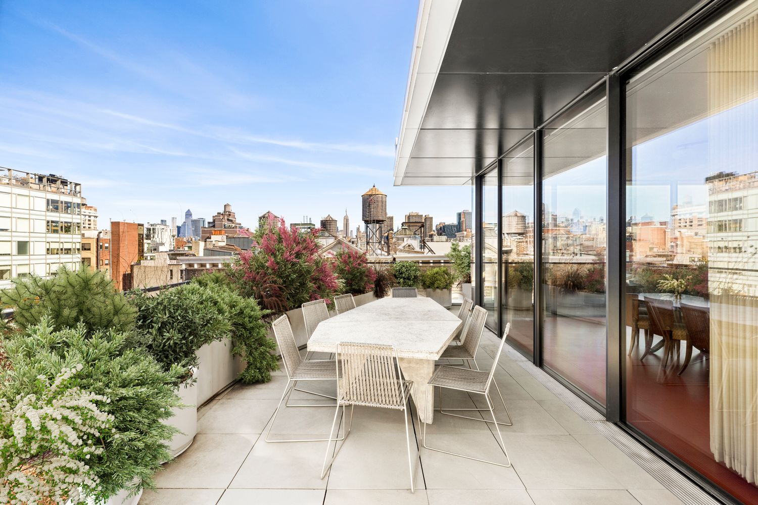 Balcony with outdoor dining table and chairs, city skyline in background, potted plants, glass sliding door, clear sky.