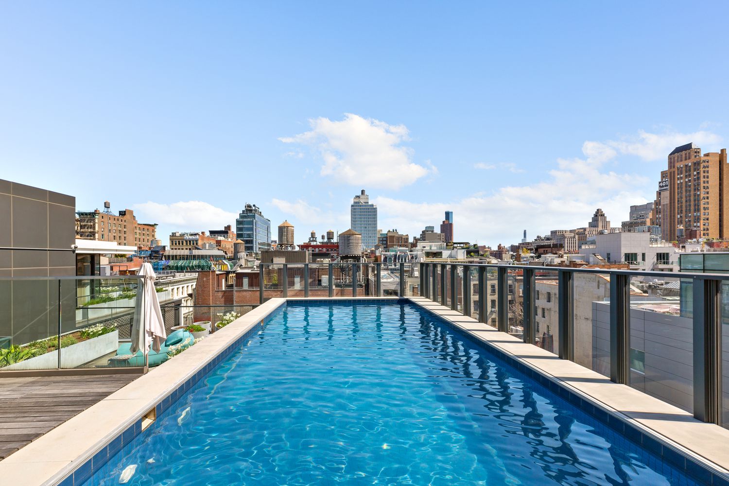 Rooftop swimming pool with city skyline in the background, blue sky with clouds, glass railing, and poolside umbrellas.