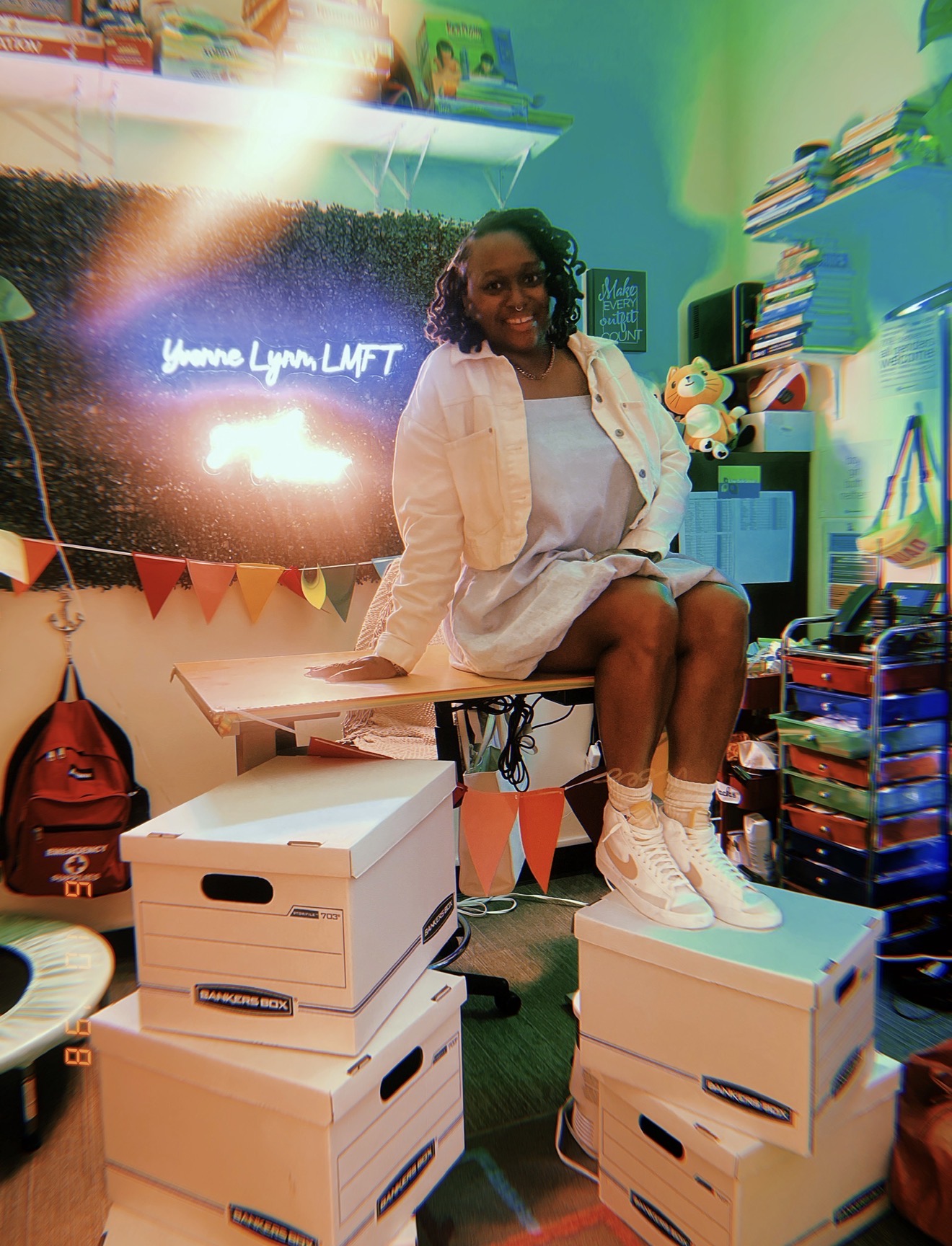 Person sitting on stacked boxes in a colorful room with books, a TV, and decorations, smiling at the camera.