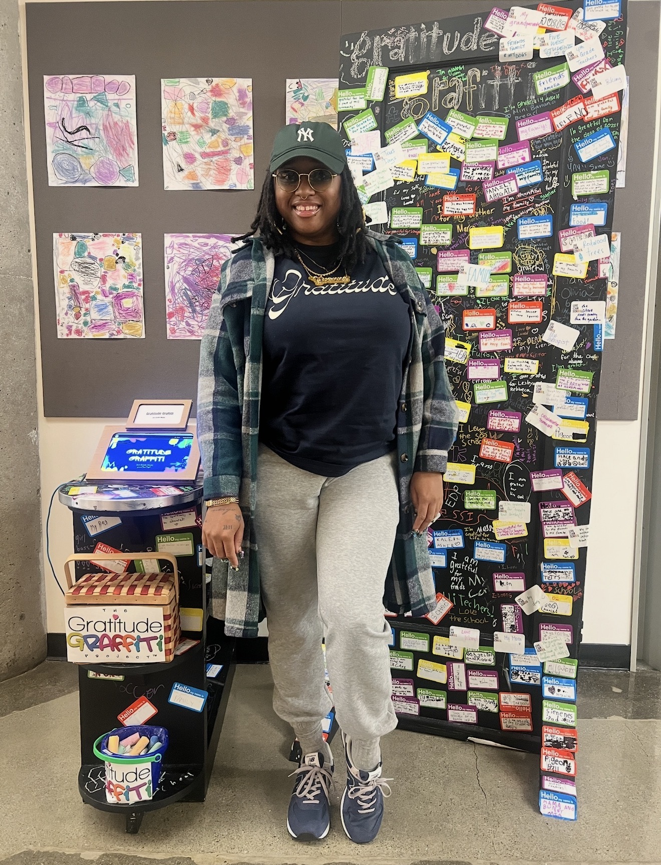 Woman standing in front of a gratitude wall with colorful notes, wearing a plaid shirt, dark hoodie, gray sweatpants, and sneakers.