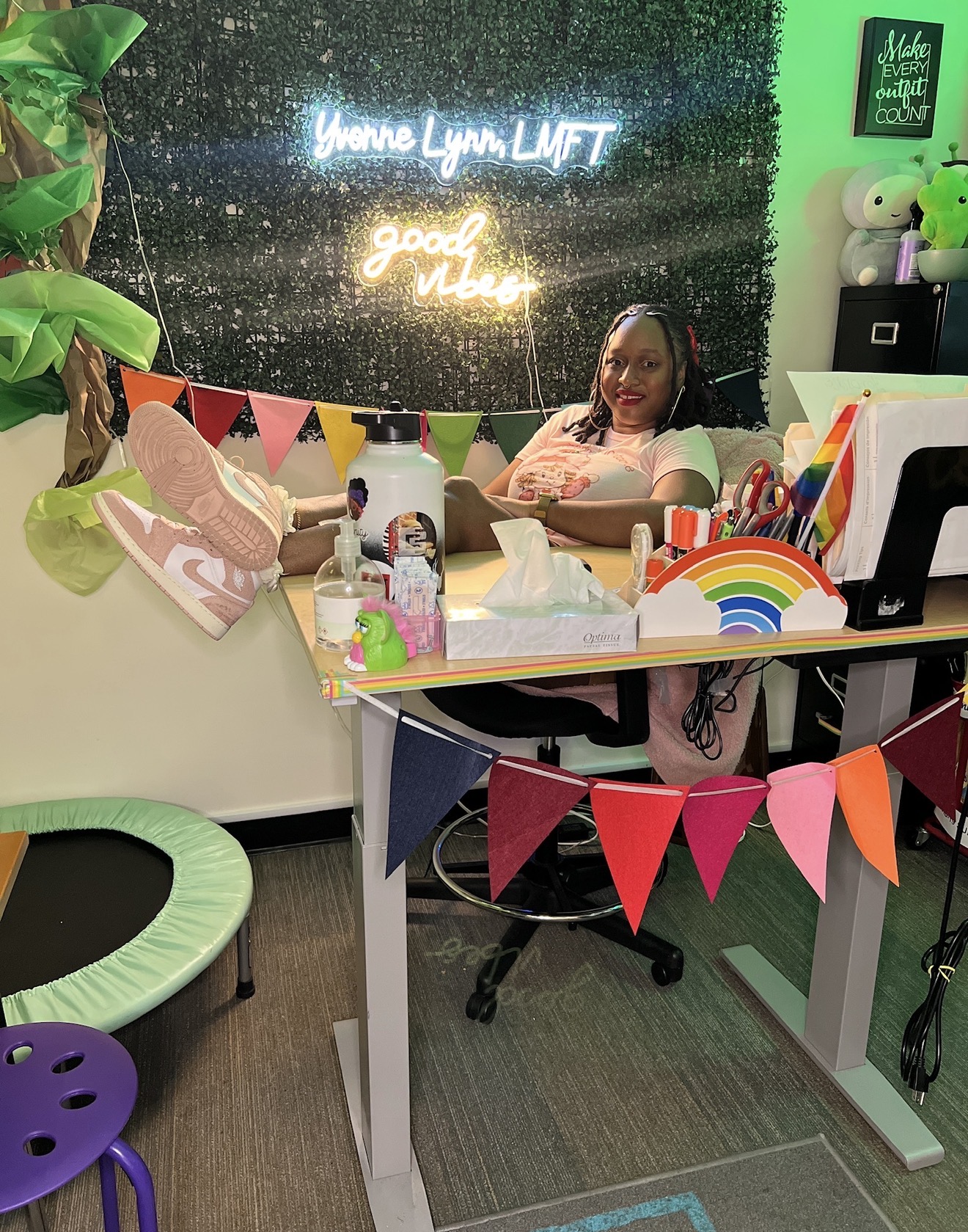 Woman sitting at a desk with decorations, neon signs, and a rainbow, in a colorful room with plants and banners.
