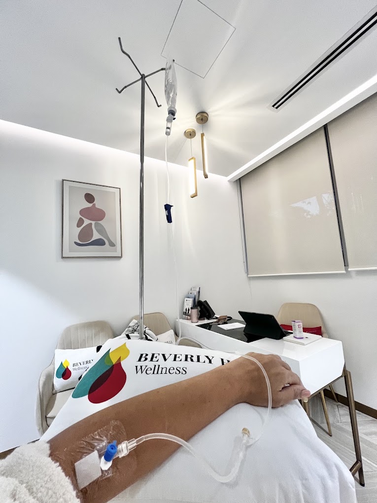 Hospital room with IV drip, patient arm, table, chairs, and abstract artwork on wall.