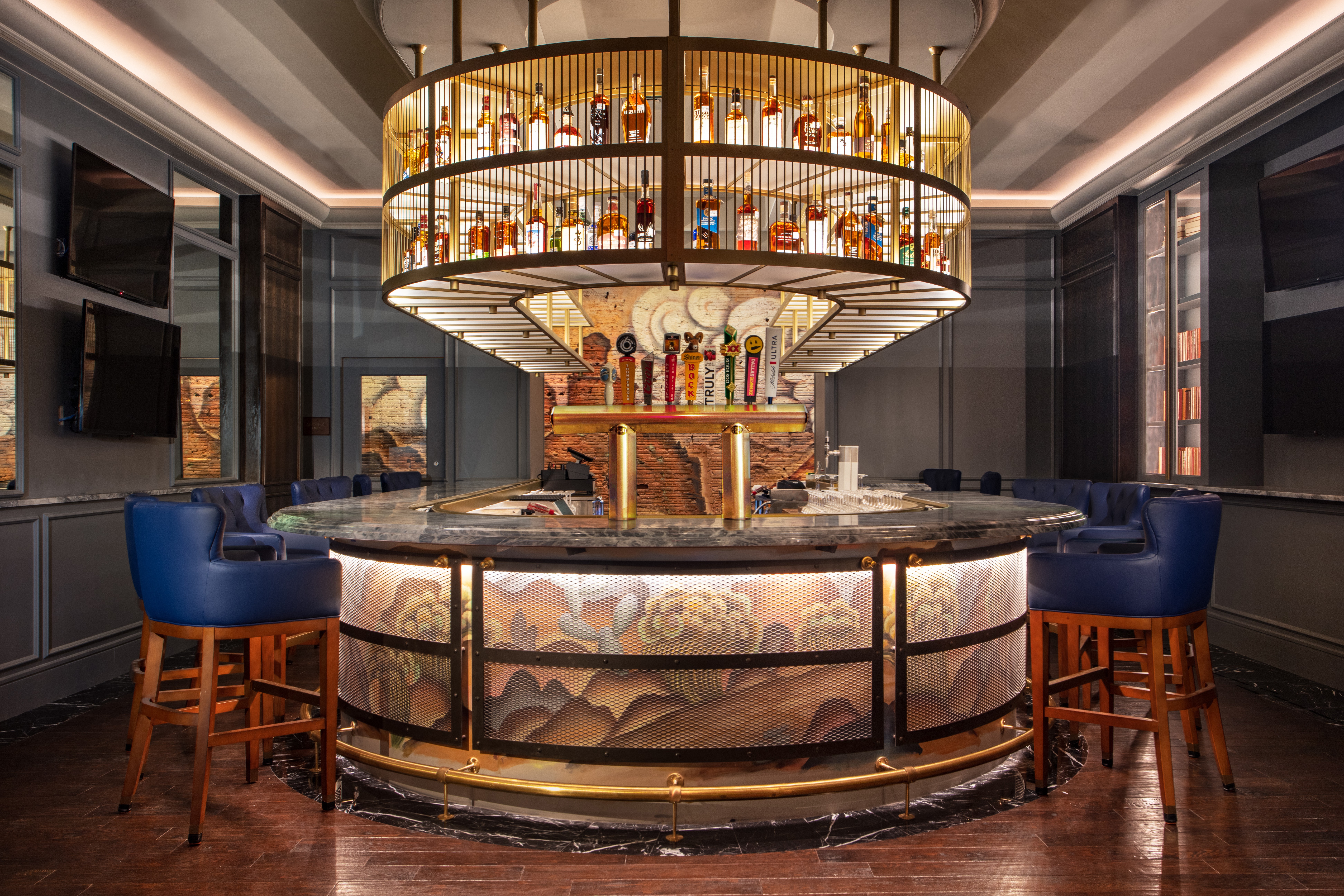 Bar with a circular counter, blue chairs, and a large, multi-tiered illuminated shelving unit with bottles, in a modern setting.
