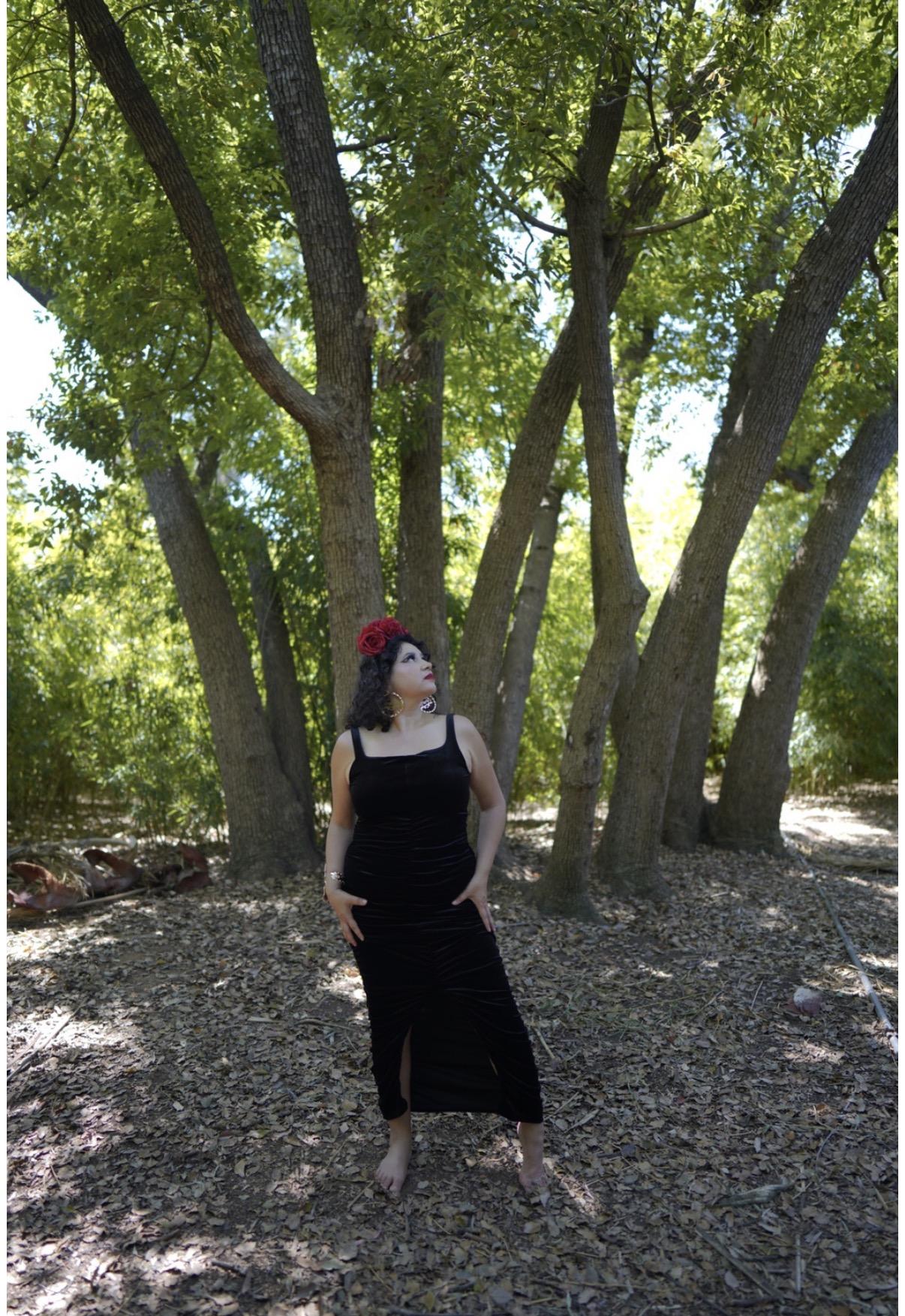Woman in black dress and red hat standing outdoors among trees with green leaves, sunlight filtering through, ground covered with leaves.