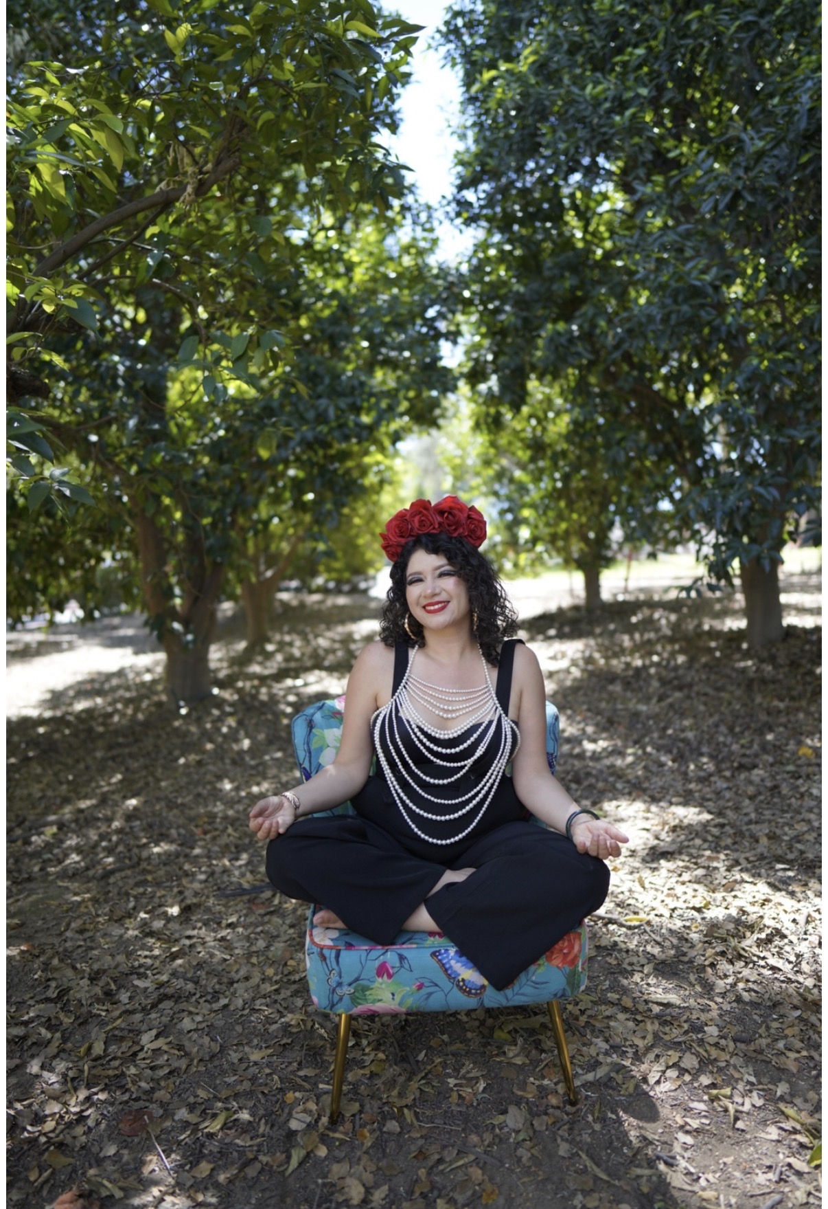 Woman sitting cross-legged on a colorful chair outdoors, wearing a flower crown and layered necklaces, surrounded by trees.