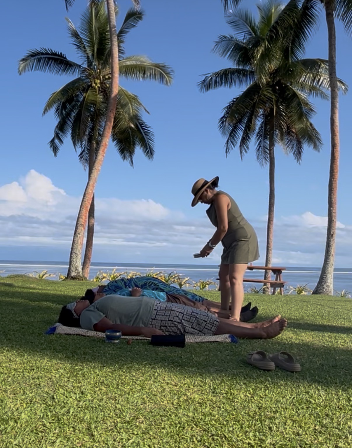Group of people lying on grass with a woman standing nearby, palm trees, blue sky, and ocean in background.