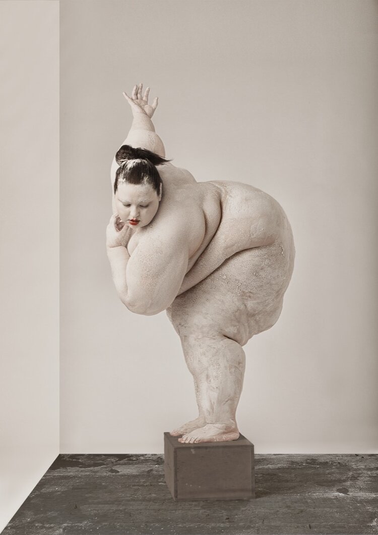 A sculpture of a person with exaggerated body features, standing on a pedestal in a gallery space.
