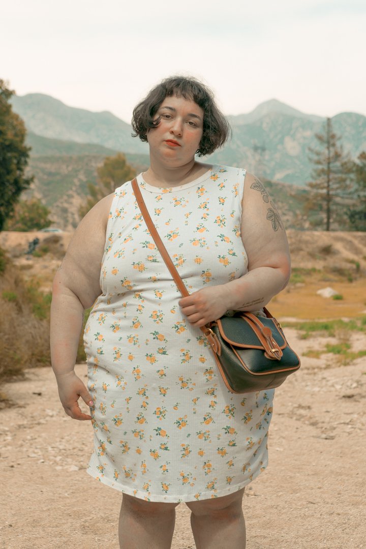 Woman with short dark hair wearing a white dress with yellow floral pattern, standing outdoors with mountains in background.