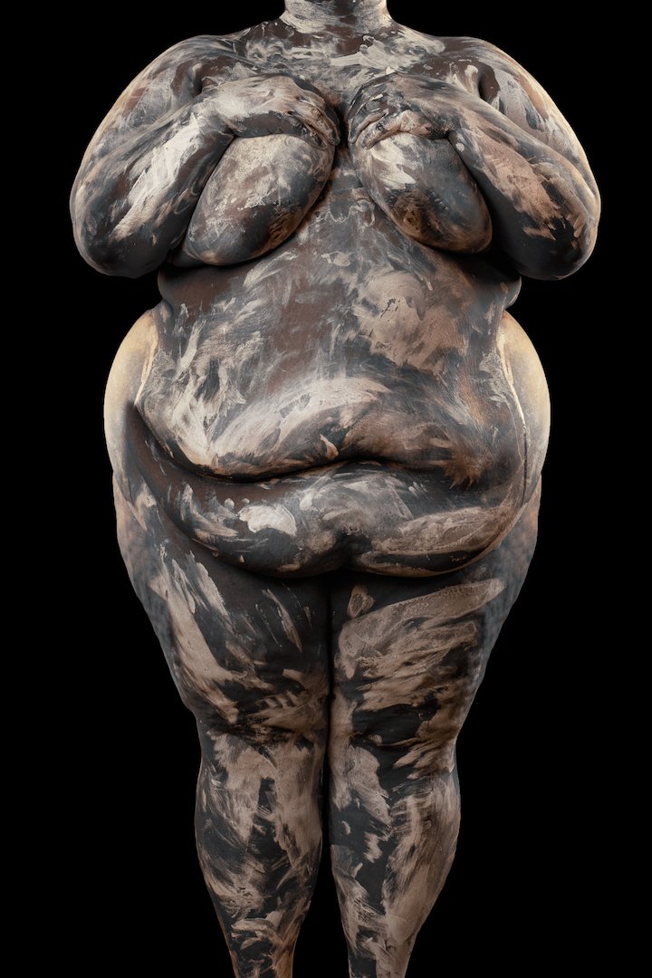 Sculpture of a nude, full-figured woman with hands covering her breasts, standing against a black background.