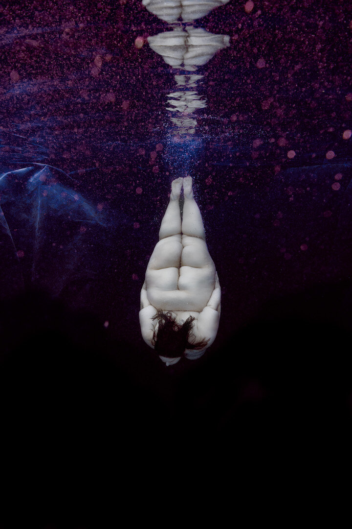 Person submerged upside down in water, with reflection visible, surrounded by dark and purple hues.