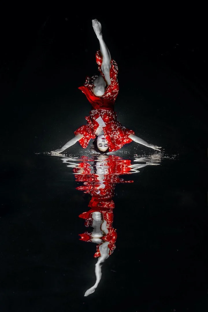 A person in a red dress performs a handstand on water, with their reflection visible below against a black background.