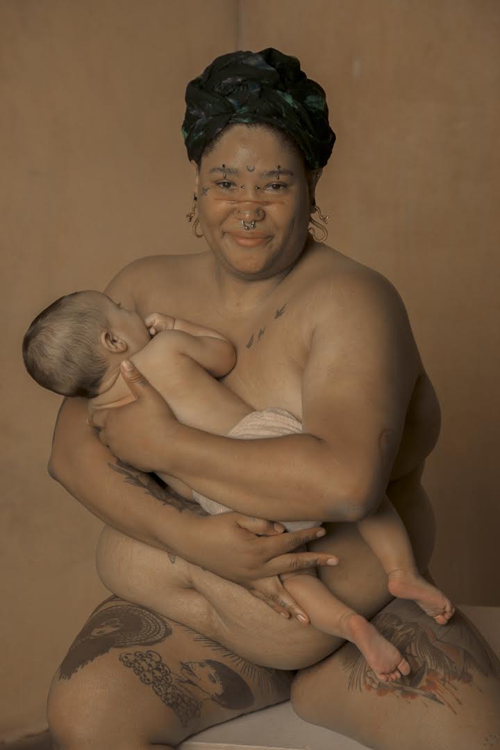 Woman holding a baby, both sitting topless against a plain background.