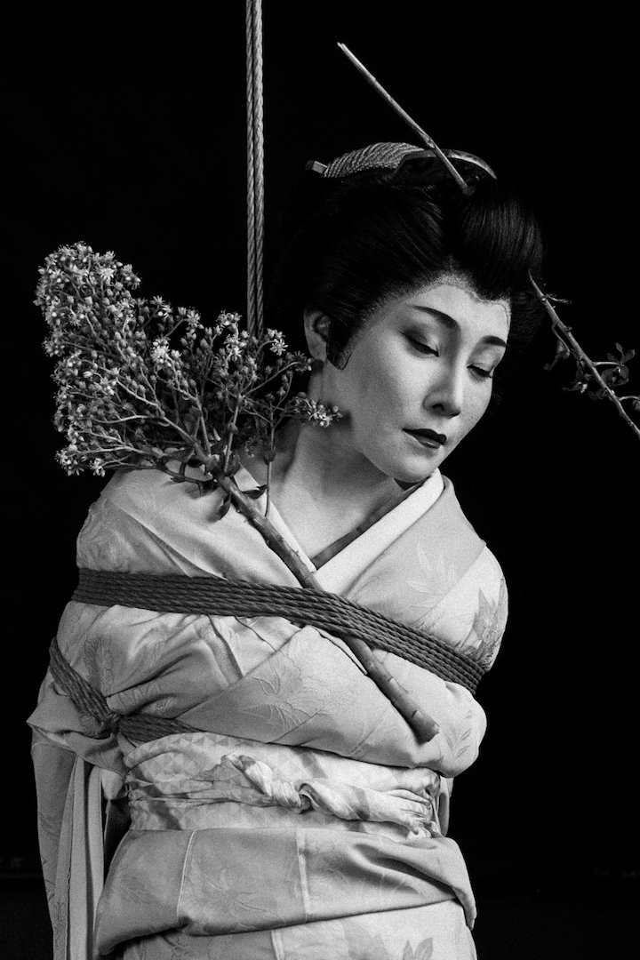 Woman in traditional attire with hairstyle and accessories, holding a bouquet of flowers, against a dark background.