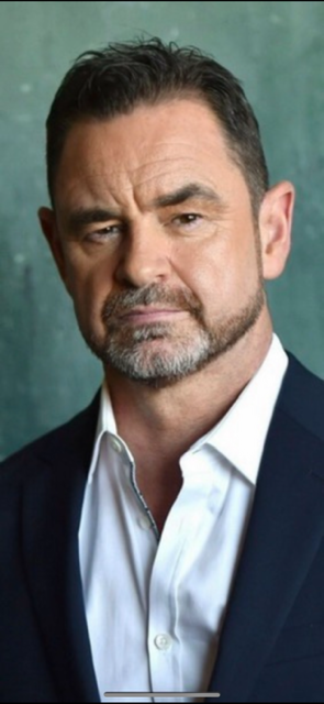 Man with dark hair, beard, wearing a white shirt and dark blazer, looking serious, against a green background.