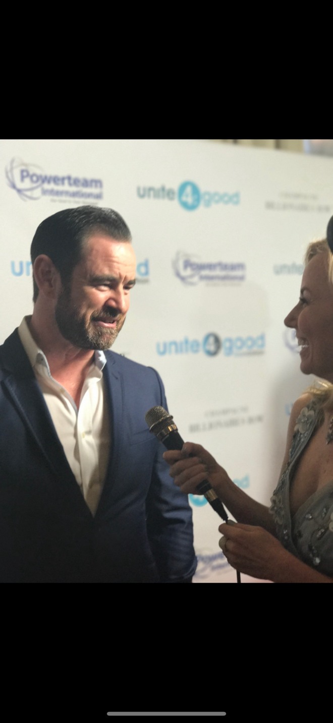 Man in a suit being interviewed by a woman holding a microphone at an event with a backdrop.