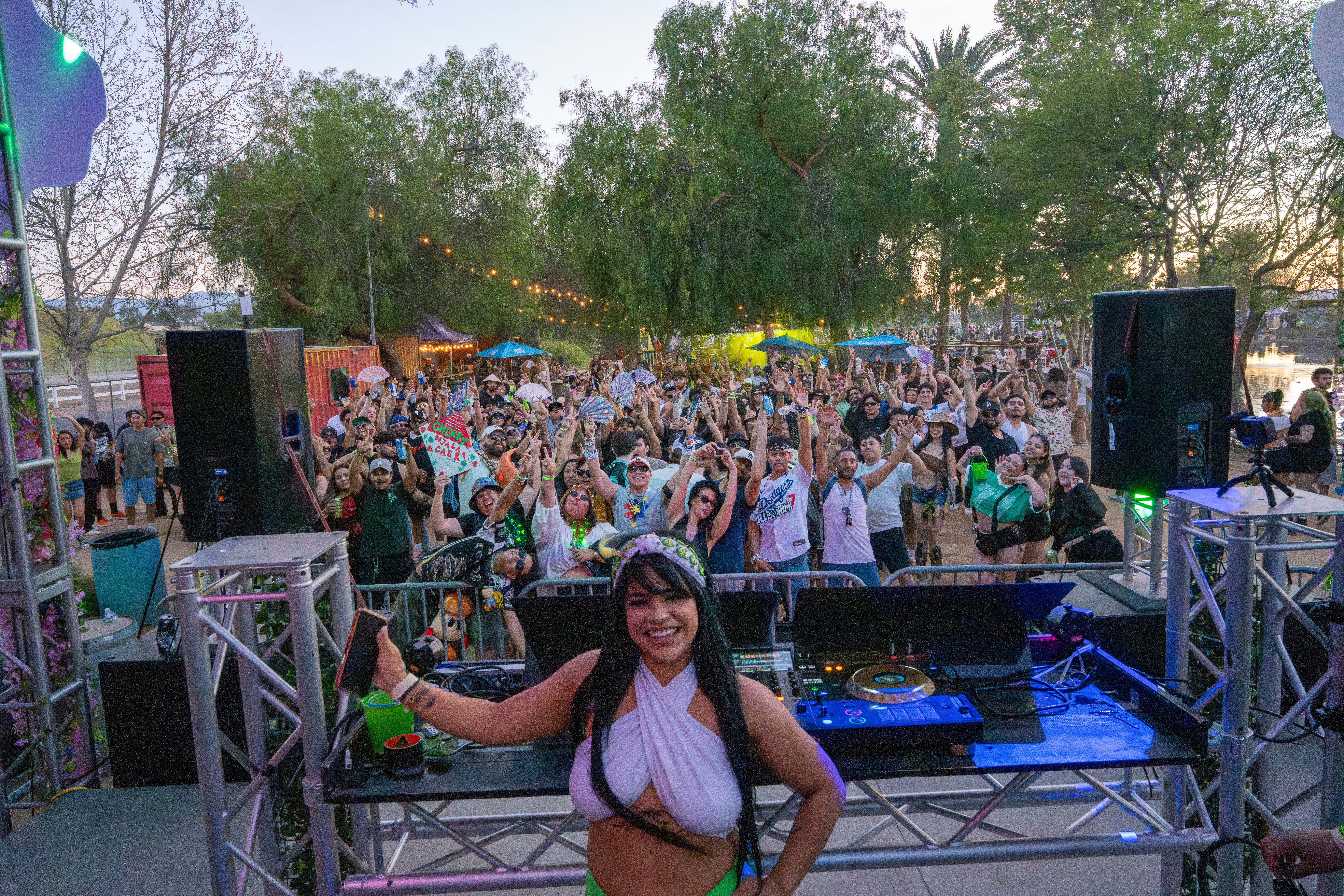 A woman stands at a DJ booth with a crowd dancing outdoors behind her, trees and sky visible in the background.