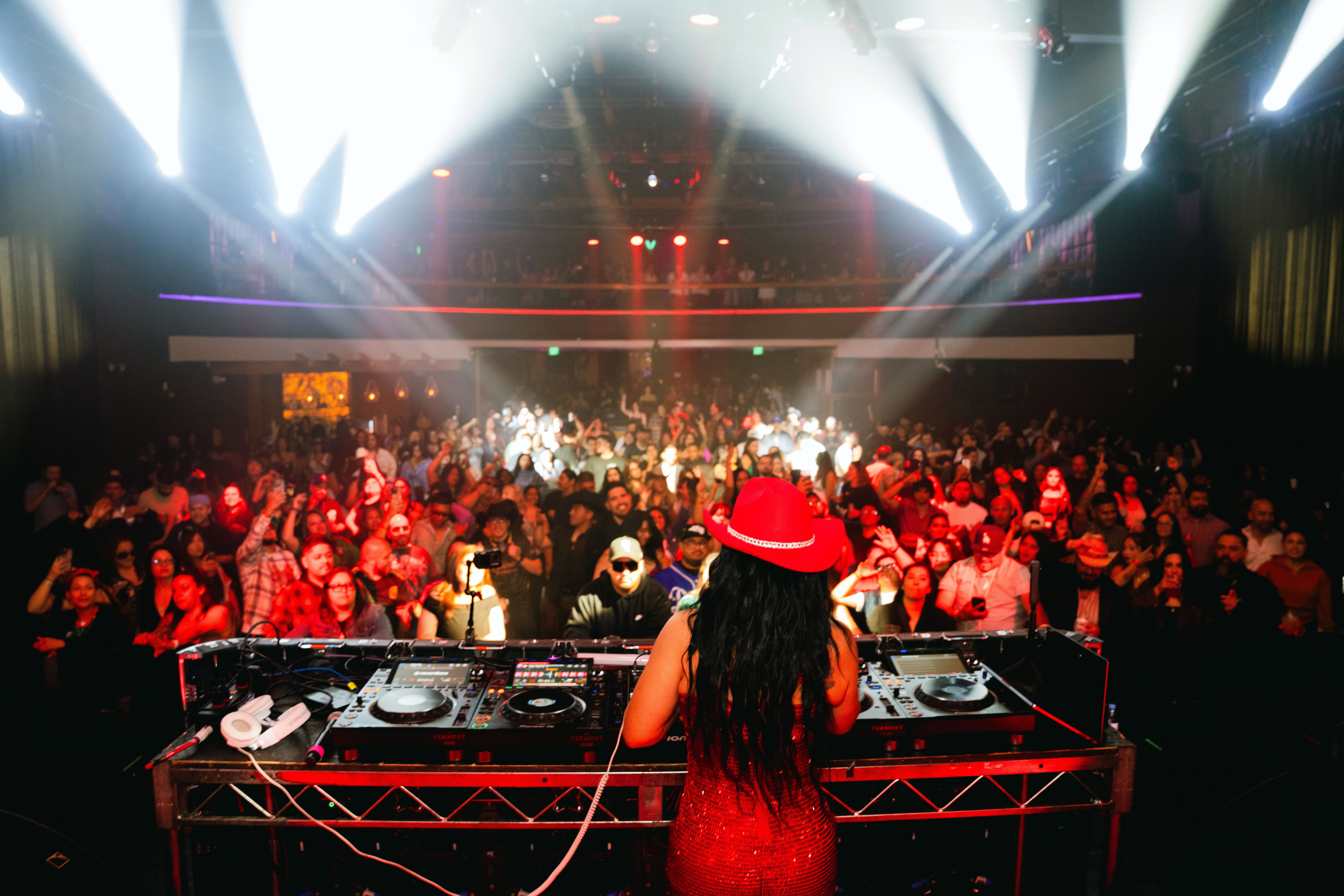 DJ performing at a crowded nightclub with colorful lights and a large audience dancing, seen from behind the DJ booth.