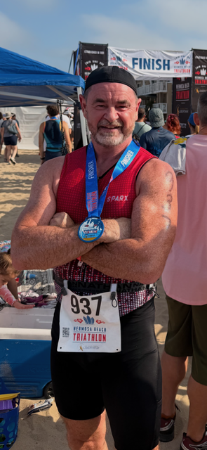 Smiling man with a beard, wearing a black cap, medal, and race bib, crossing a finish line at a race event.