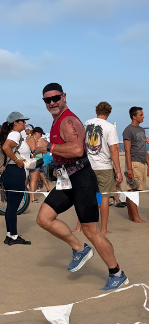 Man running on beach, wearing sunglasses, red sleeveless shirt, black shorts, and blue shoes, smiling.
