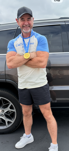 Smiling man with a medal, arms crossed, standing outdoors next to a black vehicle, wearing a cap, blue and white shirt, shorts, and sneakers.