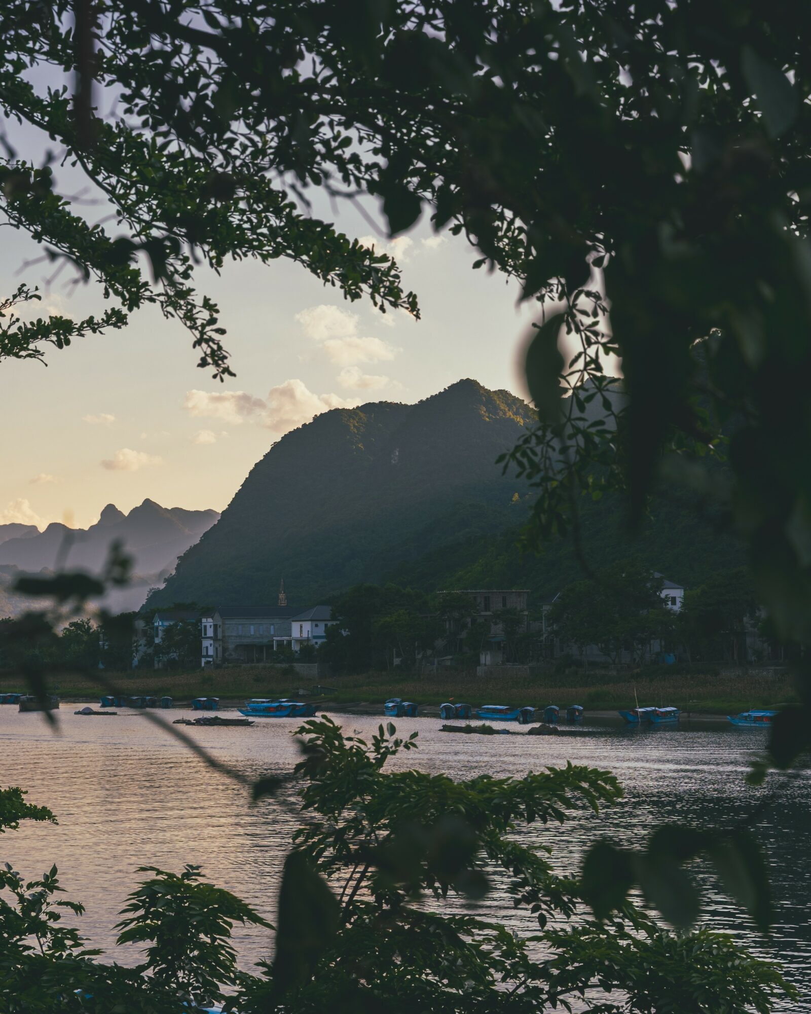 River with boats, mountain in background, trees in foreground, cloudy sky, buildings along riverbank