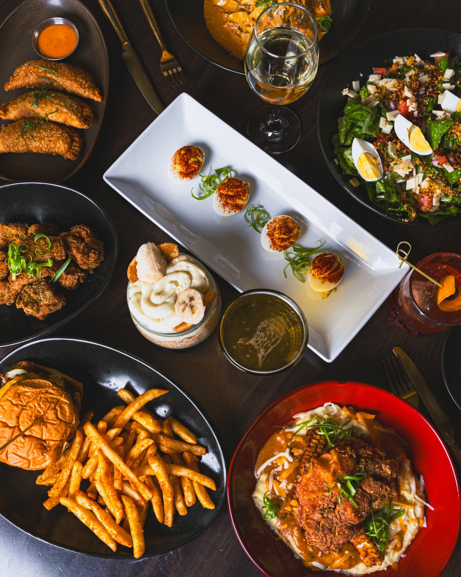Assorted dishes including fried chicken, fries, salad, and appetizers on a dark table with drinks.