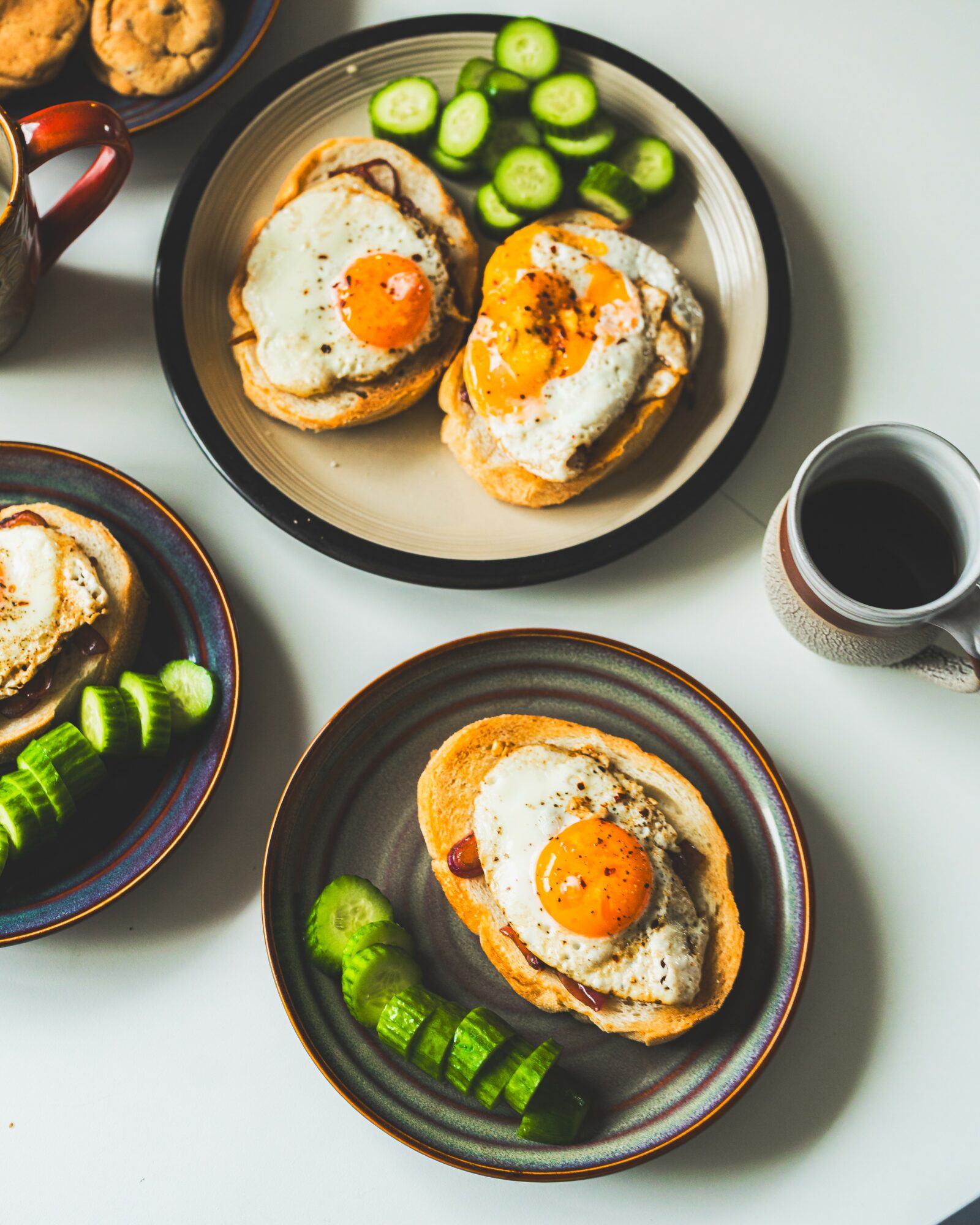Two plates with baked eggs on bread and sliced cucumbers, a cup of coffee, and a bowl with more eggs and cucumbers.