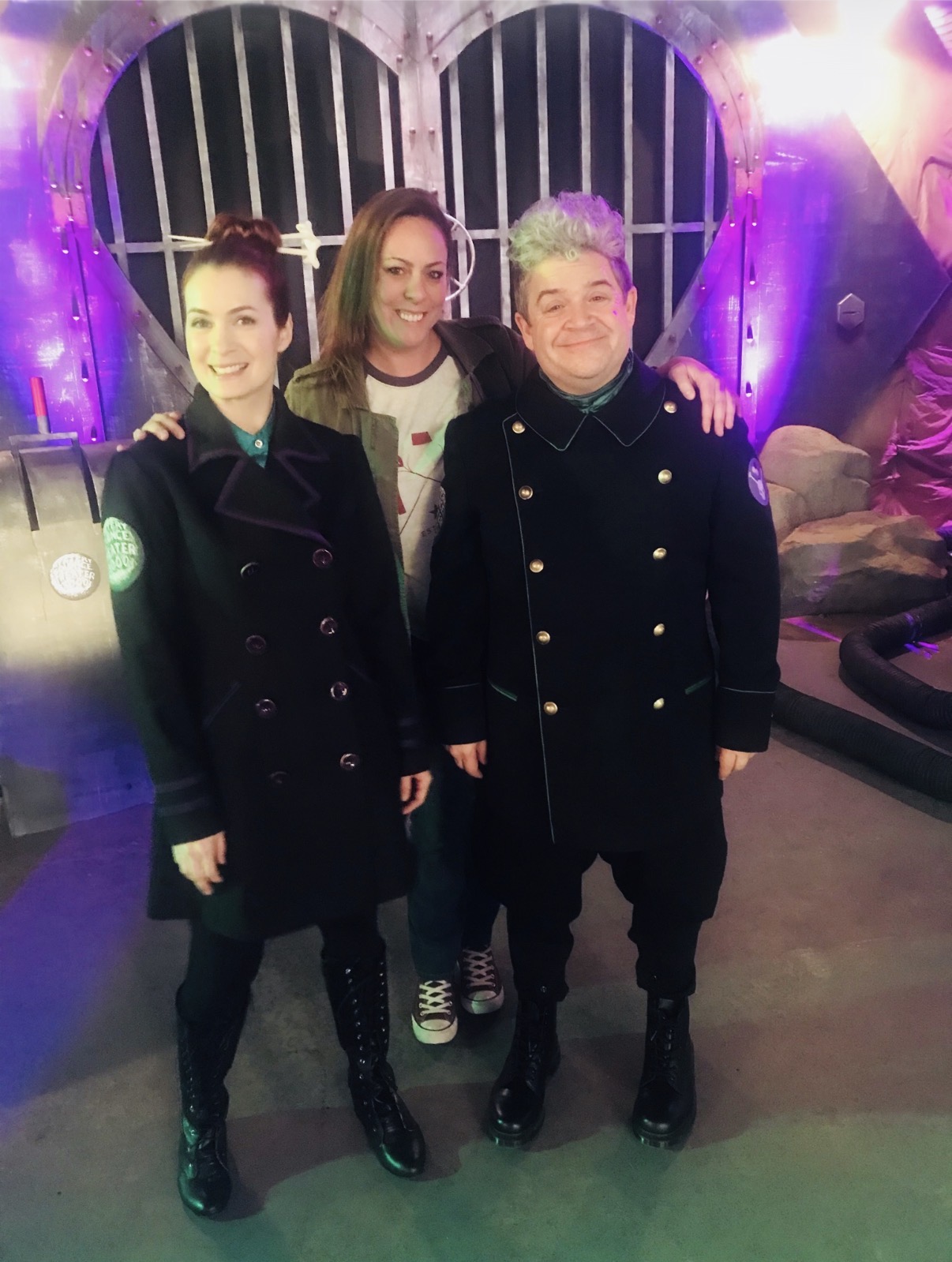 Three people stand together indoors, smiling, with colorful lighting and a decorative background behind them.